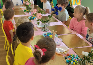 Dzieci siedzą przy stole i biorą udział w kolorowych zajęciach plastycznych. Na stole leżą pompony i druciki kreatywne. Obok dzieci stoją dwie osoby dorosłe, jedna z nich przebrana jest za lizaka.