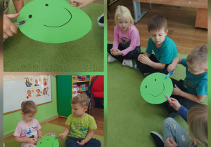 Dzieci siedzą na zielonym dywanie w sali przedszkolnej, trzymając duży zielony karton w kształcie koła z narysowaną uśmiechniętą buźką i przypinają do niego spinacze