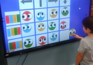 Dziecko wskazuje ikonę na ekranie dotykowym z kolorową planszą do kodowania ze strzałkami i okrągłymi symbolami.