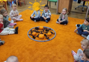 Grupa dzieci siedzi w kręgu na pomarańczowym dywanie, na środku koło z dyniami i kasztanami do zabawy i nauki kodowania.