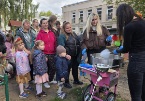 Dzieci wraz z rodzicami ustawiają się przy stanowisku z watą cukrową podczas Balu Jesieni w ogrodzie przedszkolnym. W tle widoczne są dekoracje i budynek przedszkola.