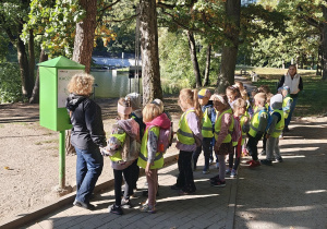 Przedszkolaki zatrzymują się przy tablicy edukacyjnej nad stawem w Arturówku, poznając ciekawostki dotyczące przyrody i zwierząt zamieszkujących okolicę.